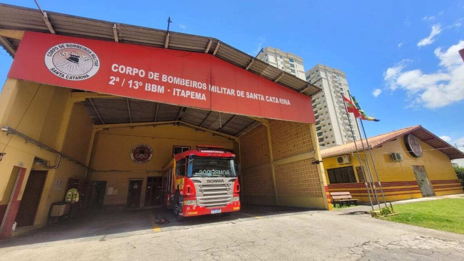 Fachada do quartel do 2ª/13º Batalhão de Bombeiros Militar de Itapema, com letreiro vermelho no alto, caminhão de bombeiros vermelho estacionado sob a cobertura e prédios altos ao fundo, em dia de céu azul.