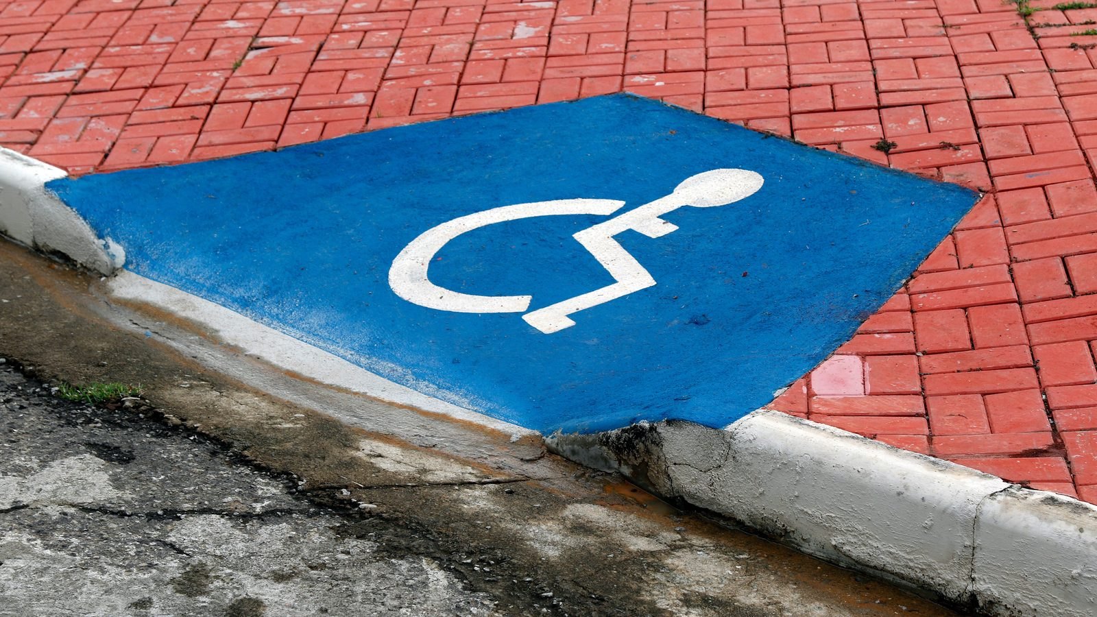 Rebaixamento de meio-fio pintado de azul com o símbolo branco de acessibilidade, ligando a calçada de piso vermelho ao asfalto de uma rua, sem nenhuma pessoa no quadro.