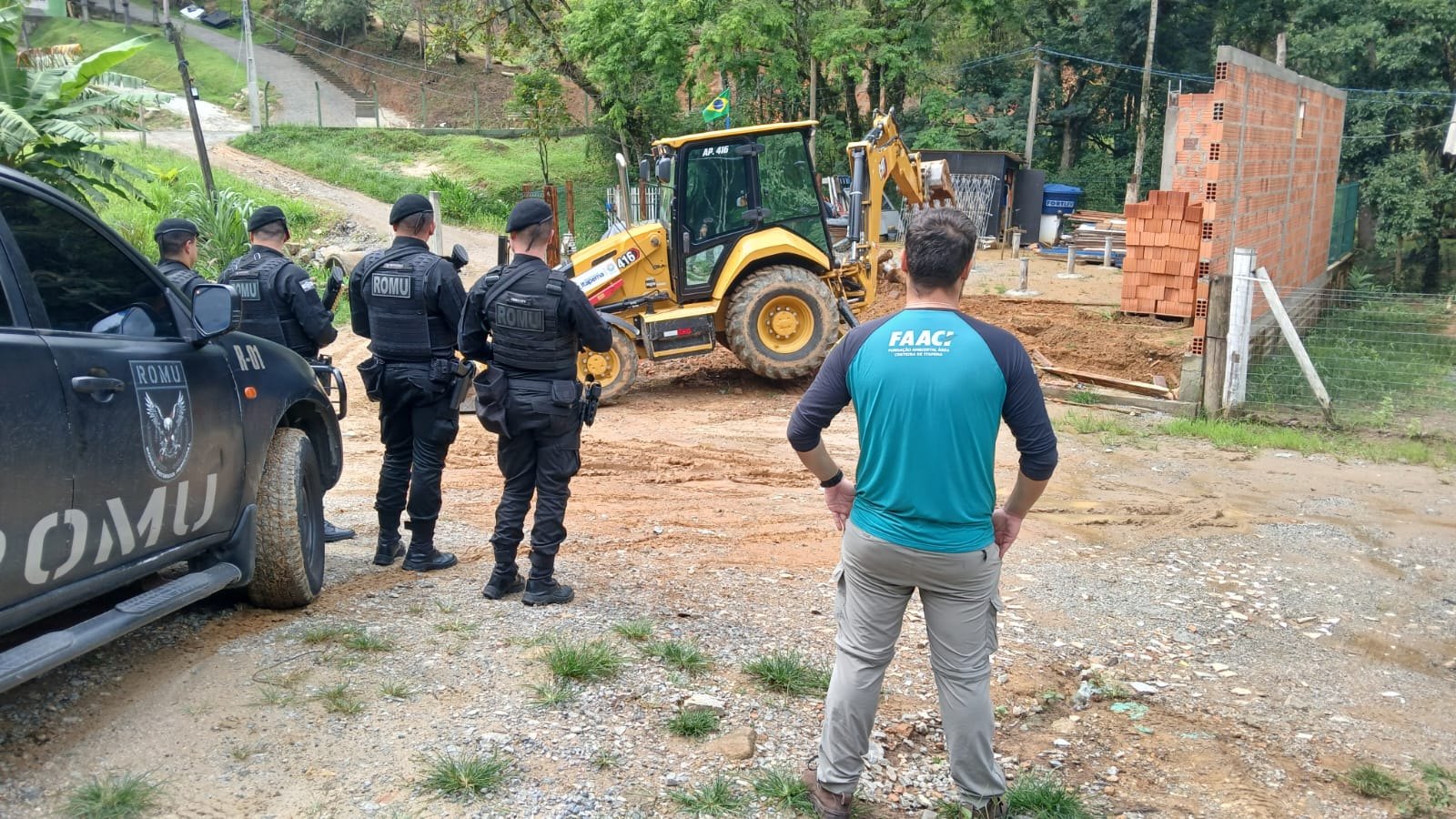 Retroescavadeira amarela parada em terreno de saibro ao lado de uma construção de tijolo sem reboco, com agentes da Guarda Municipal de costas junto a uma caminhonete e um servidor de camiseta da FAACI observando, mata fechada ao fundo.