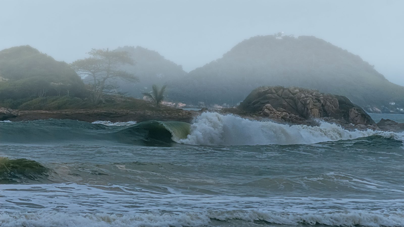 Onda grande quebrando perto da areia num dia de neblina, com espuma branca subindo à frente de um costão de pedras e morros verdes encobertos pela bruma ao fundo.
