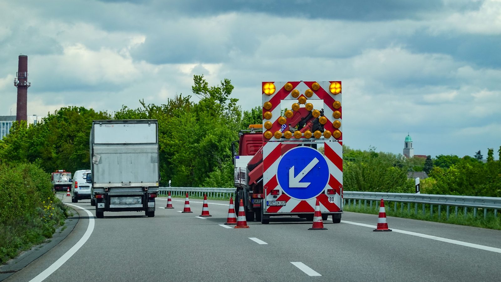 Rodovia de pista dupla com uma faixa fechada por cones laranja e brancos, um caminhão de sinalização com seta luminosa apontando para a esquerda e um caminhão baú seguindo pela faixa liberada, sob céu nublado.