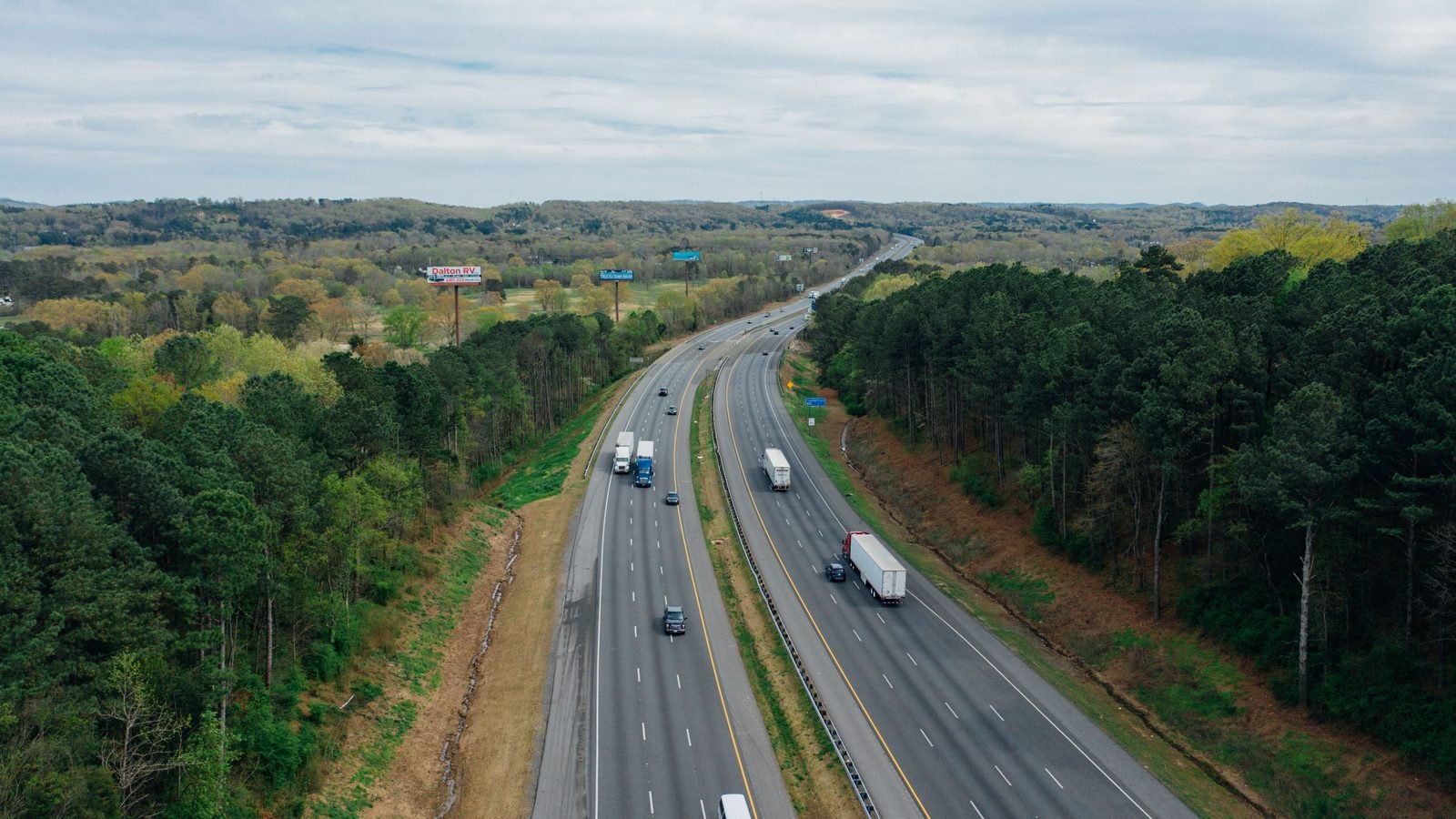 Vista aérea de uma rodovia de pista dupla cortando uma área de mata, com caminhões de carga e carros circulando nos dois sentidos e o canteiro central separando as faixas.