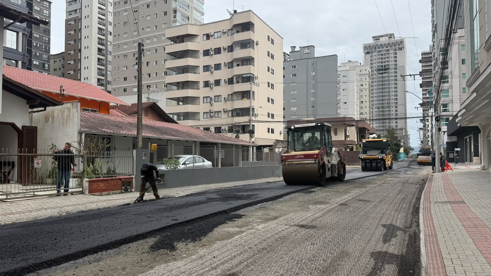 Rolo compactador passando sobre asfalto novo e escuro na Rua 282, na Meia Praia, em Itapema, com um operário espalhando massa asfáltica com um rodo à esquerda, um segundo rolo mais ao fundo, calçada de pavers vermelhos à direita e prédios altos ao longo de toda a rua.