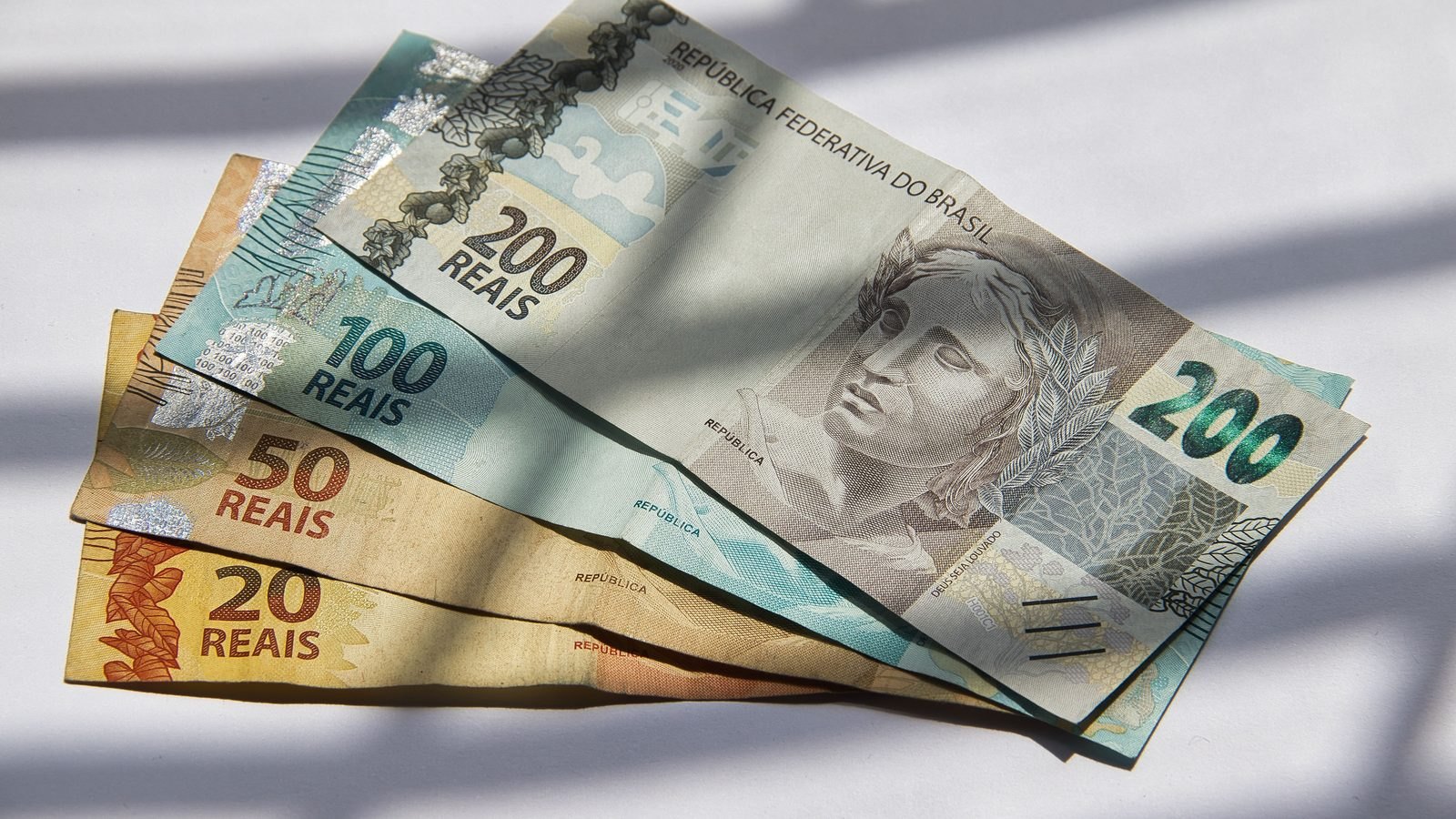 Quatro cédulas de real abertas em leque sobre uma superfície branca, com as notas de 200, 100, 50 e 20 reais aparecendo pelo canto, sob luz de sol coada por uma persiana.