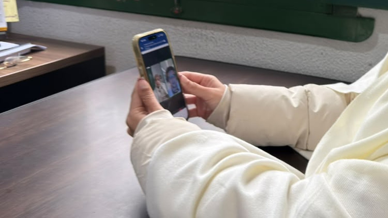 Mãos de uma pessoa sentada à mesa segurando um celular com uma consulta por vídeo aberta na tela, dentro de uma sala de paredes claras com janela de madeira verde ao fundo.