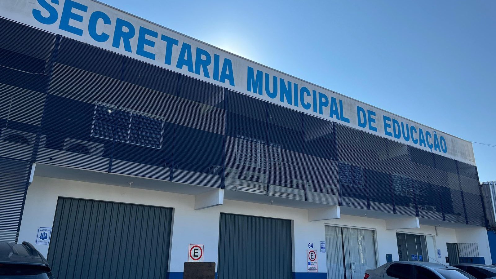 Fachada da Secretaria Municipal de Educação de Itapema, prédio de dois andares com o nome em letras azuis no alto, grades escuras no pavimento superior, portões verdes na frente e carros estacionados na calçada.