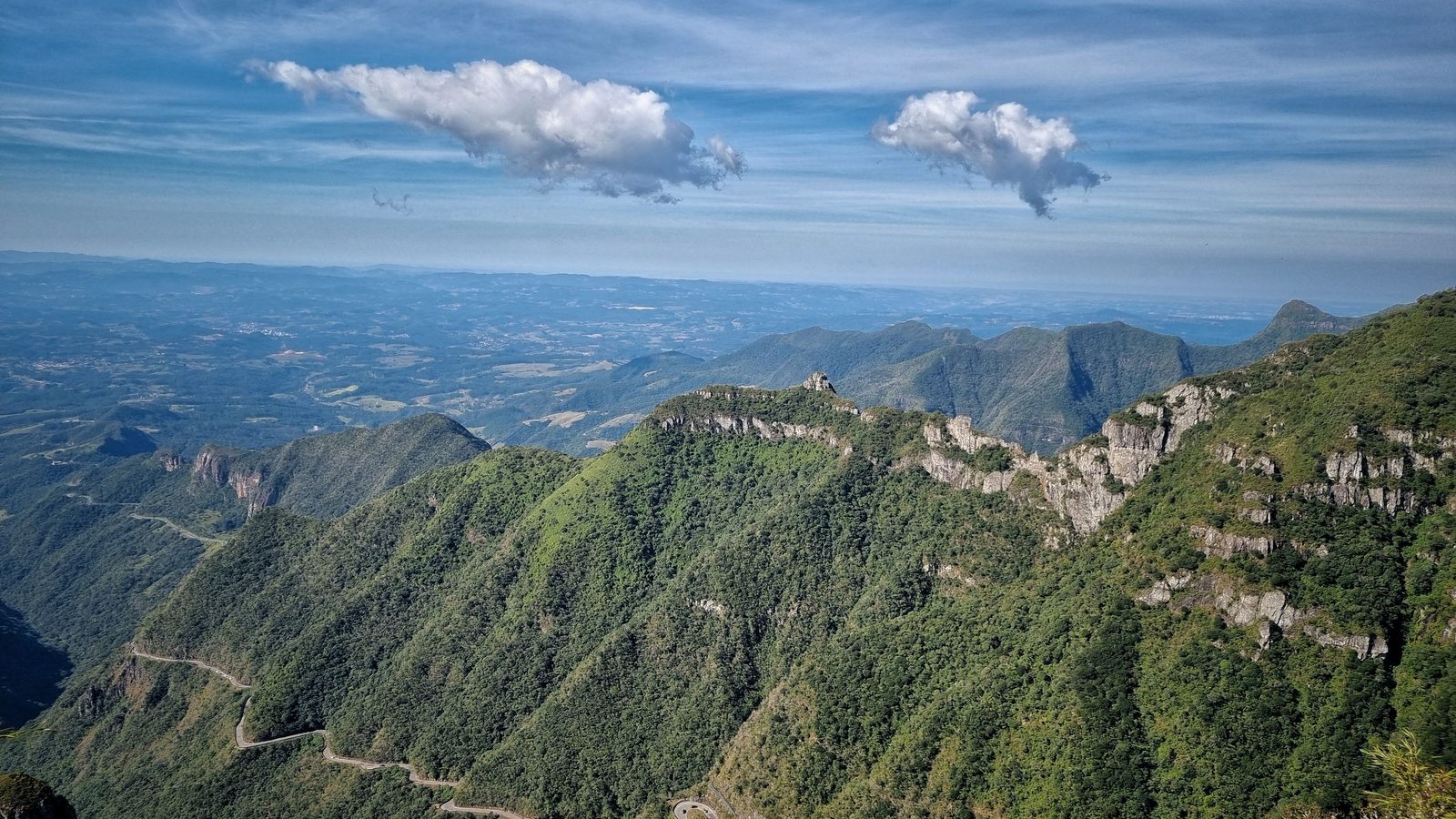 Estrada sinuosa descendo por encostas cobertas de vegetação, com montanhas se sobrepondo até o horizonte sob céu claro.