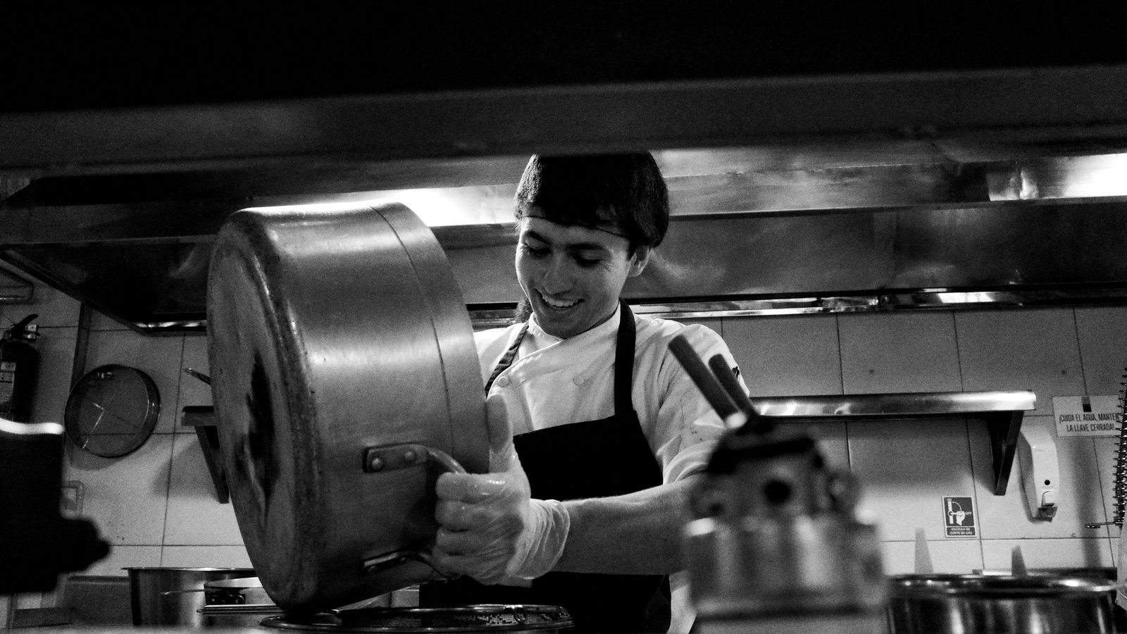 Cozinheiro de dólmã branco e avental escuro sorrindo enquanto despeja o conteúdo de uma panela grande sobre o fogão de uma cozinha profissional, em foto preto e branco.