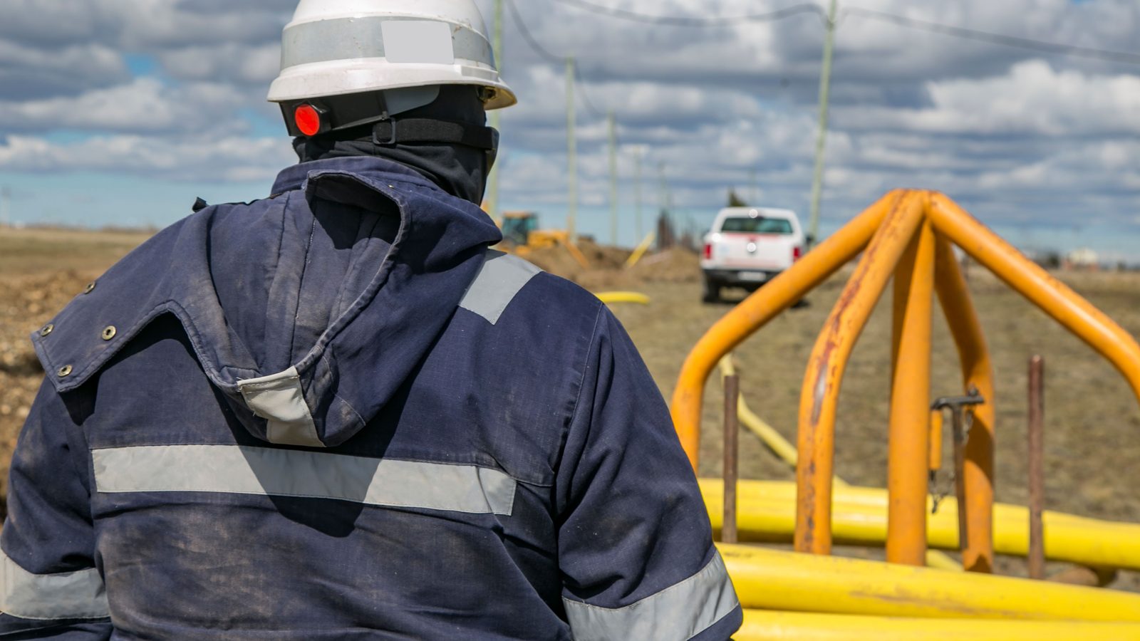 Trabalhador visto de costas em um canteiro de obras, com capacete branco, lanterna presa na frente do capacete e jaqueta azul com faixas refletivas, olhando para tubulações amarelas empilhadas no terreno, com céu nublado ao fundo.