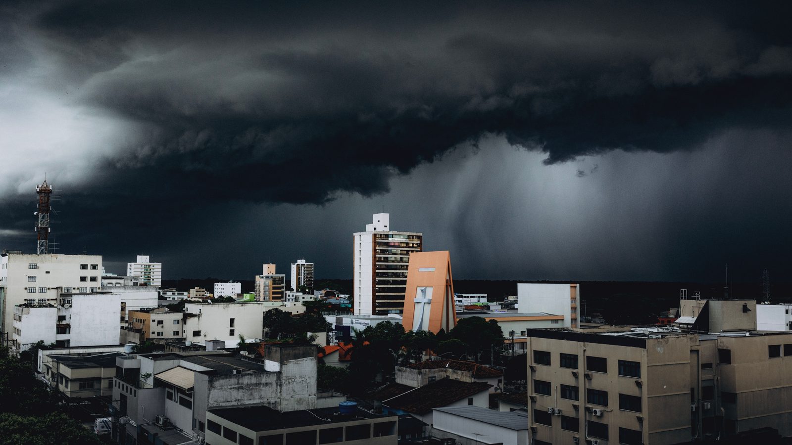 Nuvem de temporal muito escura e baixa cobrindo o céu inteiro sobre os prédios de uma cidade, com uma cortina de chuva descendo ao fundo à direita e uma torre de antena recortada contra o cinza à esquerda.