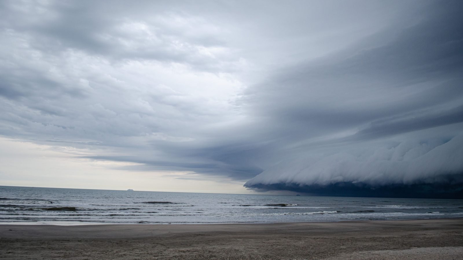 Nuvem escura e alongada de tempestade avançando sobre o mar em direção a uma faixa de areia vazia, com a água esverdeada e o céu claro ainda visível no lado esquerdo do horizonte.