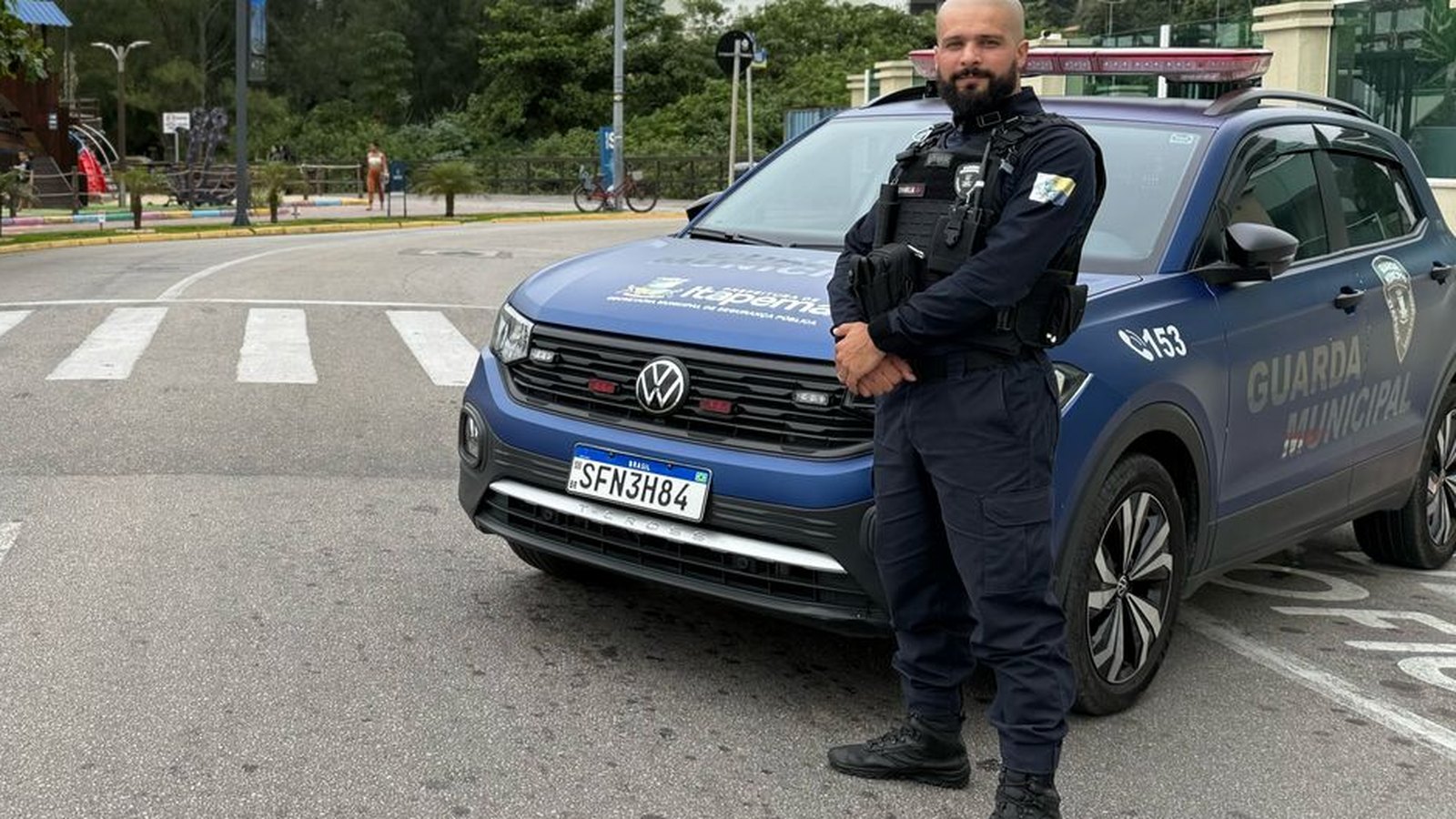 Agente da Guarda Municipal de Itapema, de uniforme escuro e colete, em pé ao lado de uma viatura azul atravessada na pista de uma avenida da orla, com faixa de pedestres à frente e a rua vazia.