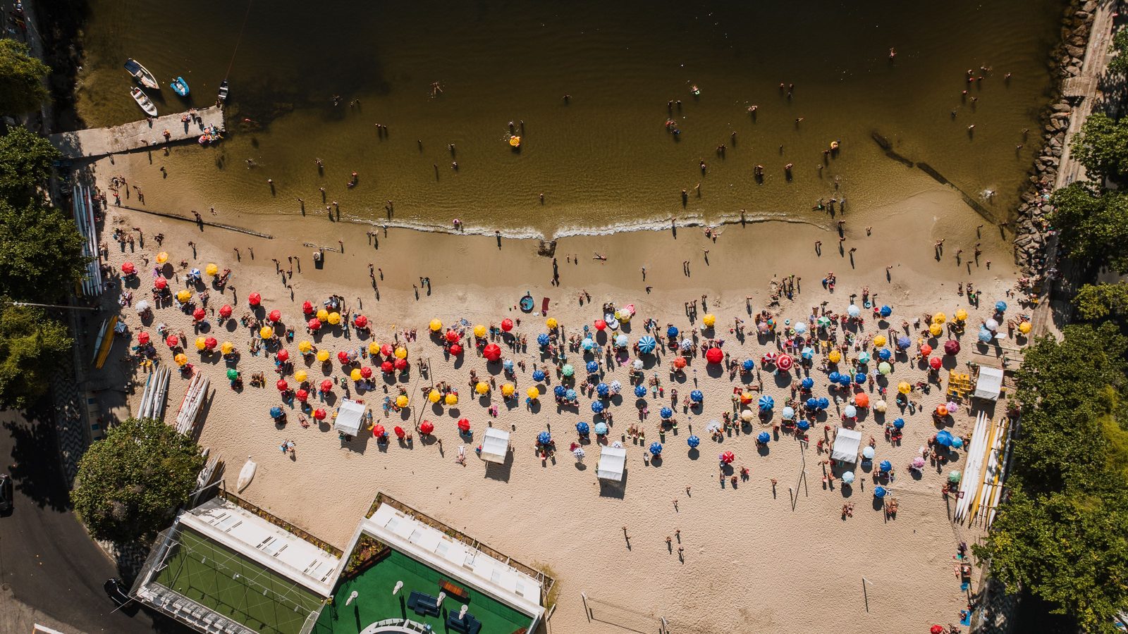 Vista de drone de uma faixa de praia cheia, com dezenas de guarda-sóis coloridos enfileirados sobre a areia clara, banhistas dentro da água rasa e uma quadra e um trapiche nas pontas do quadro.