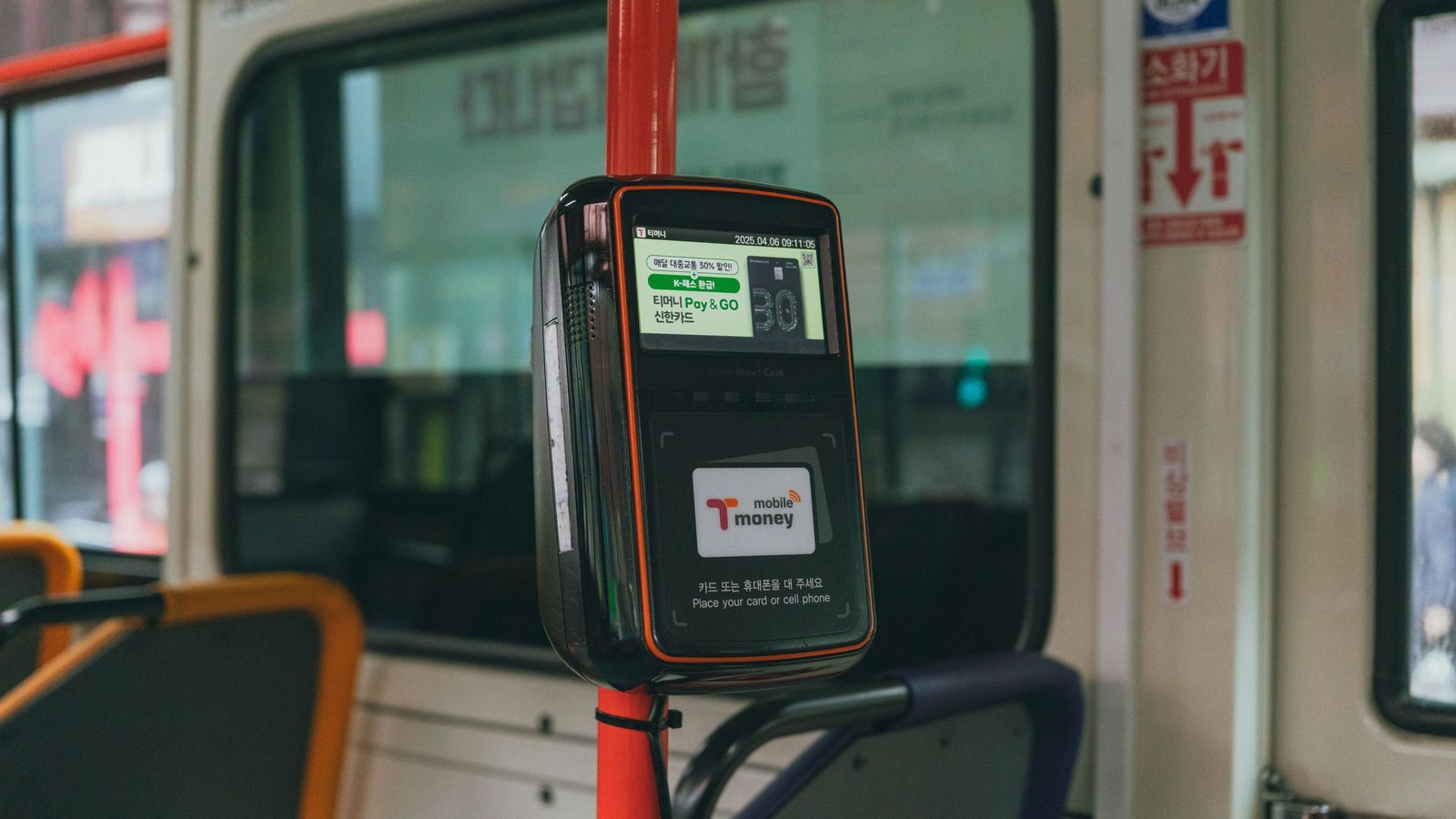 Validador de cartão preso a uma barra vermelha dentro de um ônibus urbano, com tela acesa, área de aproximação do cartão e janelas do veículo desfocadas ao fundo.