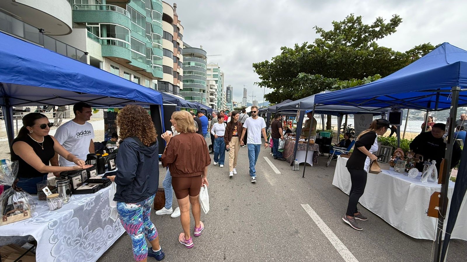 Tendas azuis montadas nos dois lados da avenida da orla de Itapema, com bancas de artesanato, visitantes circulando pelo meio da via e a linha de prédios da praia ao fundo.